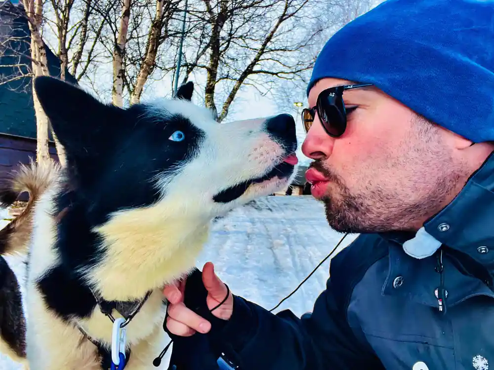 Man in blue hat kissing black and white sled dog